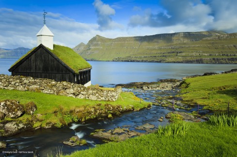A stream running into the Funningsfjordur and a traditional turf or grass roofed church, located in the picturesque village of Funningur.